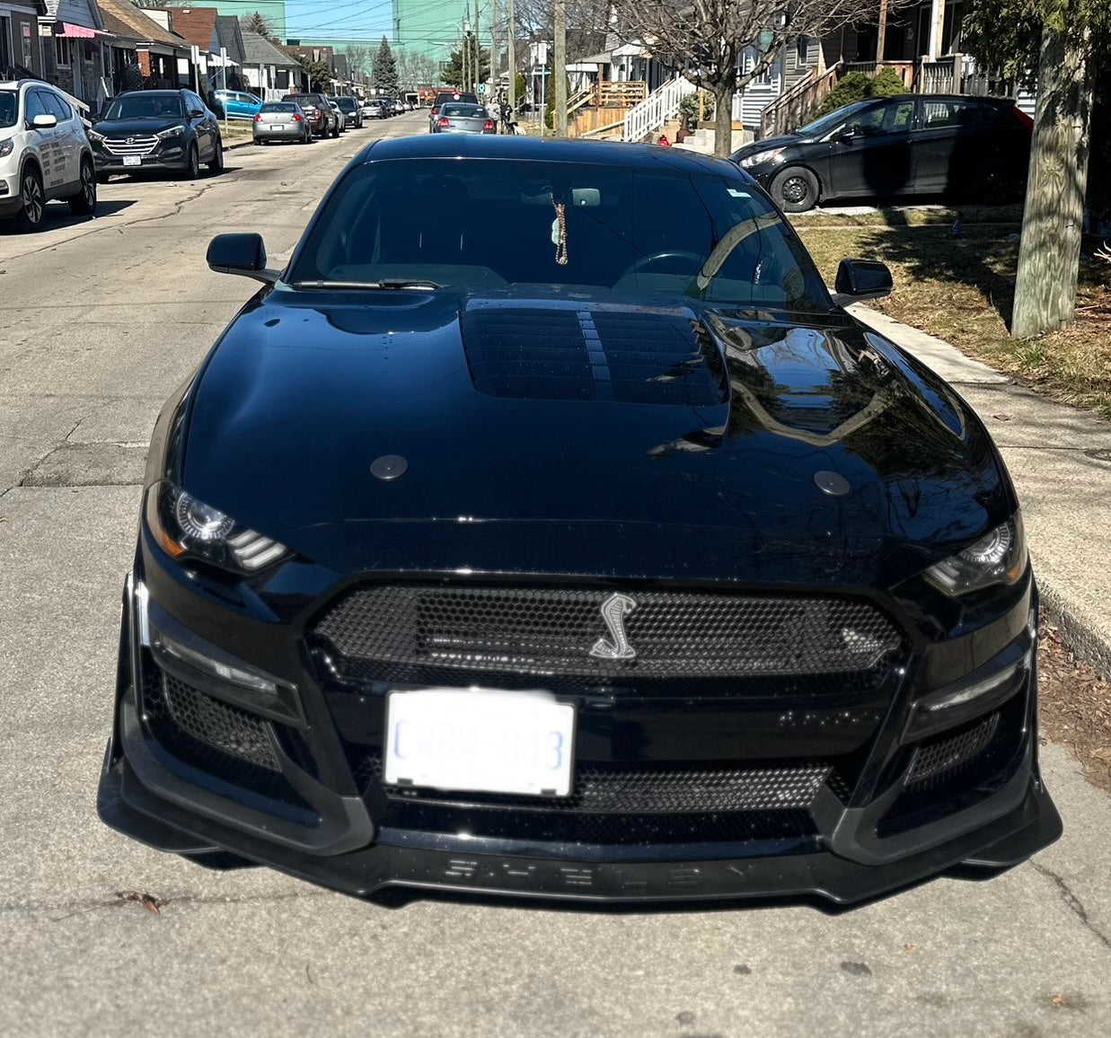 Front view of a black 2018–2022 Ford Mustang featuring a Shelby GT500 front bumper conversion kit, parked on a residential street, showcasing the aggressive grille and aerodynamic Shelby styling.
